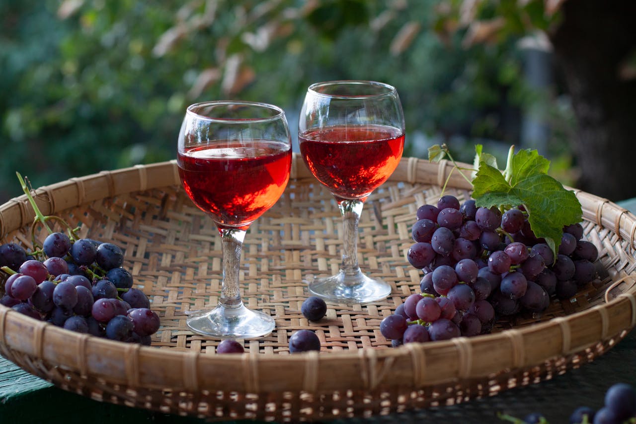 Two glasses of red wine on a wicker tray with bunches of fresh grapes under natural light.